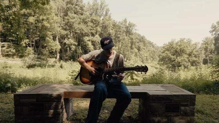 Kaila Sit playing acoustic guitar on a wooden bench in a green field